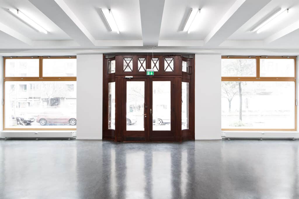 Innenansicht eines hell erleuchteten Raums mit polierten Böden und hölzernen Doppeltüren mit Glaspaneelen in der Mitte. Große Fenster auf beiden Seiten der Türen bieten einen Blick auf geparkte Autos und Bäume draußen. Deckenleuchten verstärken das moderne Ambiente des Raums.