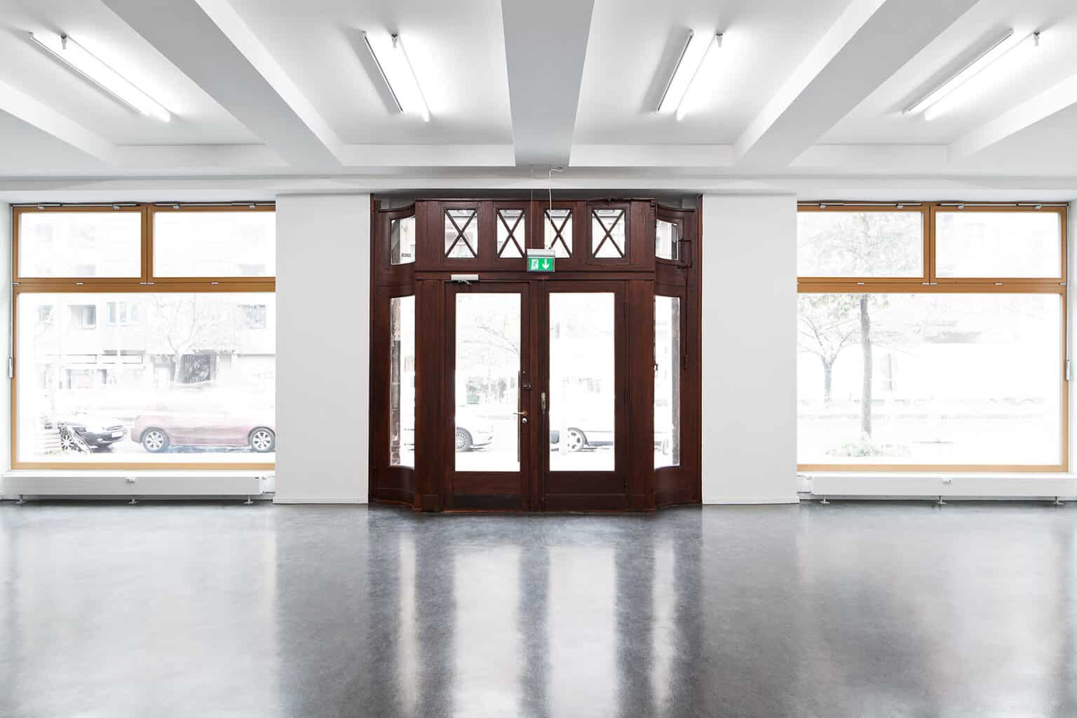 Innenansicht eines hell erleuchteten Raums mit polierten Böden und hölzernen Doppeltüren mit Glaspaneelen in der Mitte. Große Fenster auf beiden Seiten der Türen bieten einen Blick auf geparkte Autos und Bäume draußen. Deckenleuchten verstärken das moderne Ambiente des Raums.